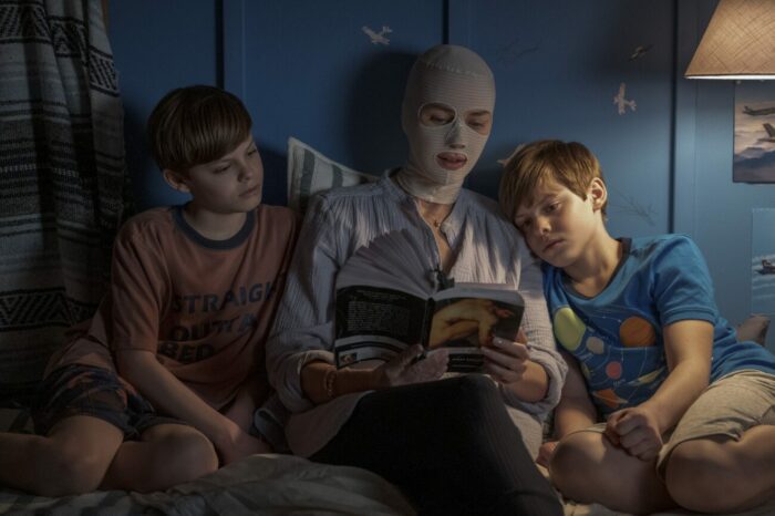 A woman in a post-surgical mask reads to two twin boys in bed.