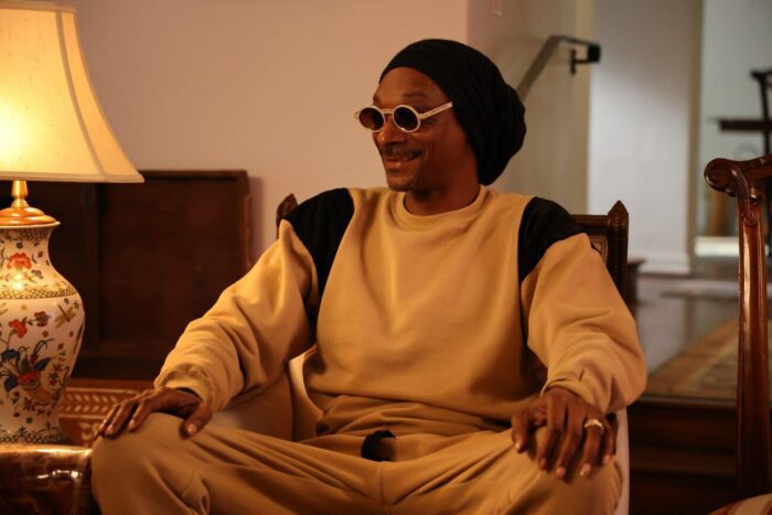 Snoop Dogg, a black man wearing a black beanie hat, circular white shades, and a tan set of a long sleeve shirt and pants with black shoulders, smiling and sitting in his living room. There is a fancy lamp to the left, illuminating the room in a soft golden glow. 