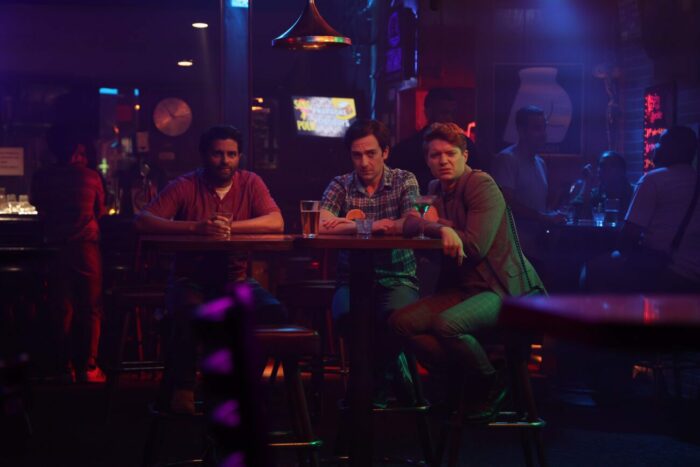 Angry Mike (Asif Ali), an Indian man with short black hair, Sid, and Runway Dave (Brendan Scannell), a white man with ginger hair sitting together at a table in a night club, several different drinks in front of them. The room is dark, lit in neon purple, blue, red, and yellow.