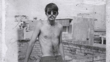 A young bare-chested man wears sunglasses and faces the camera in a black-and-white photograph from the 1970s.
