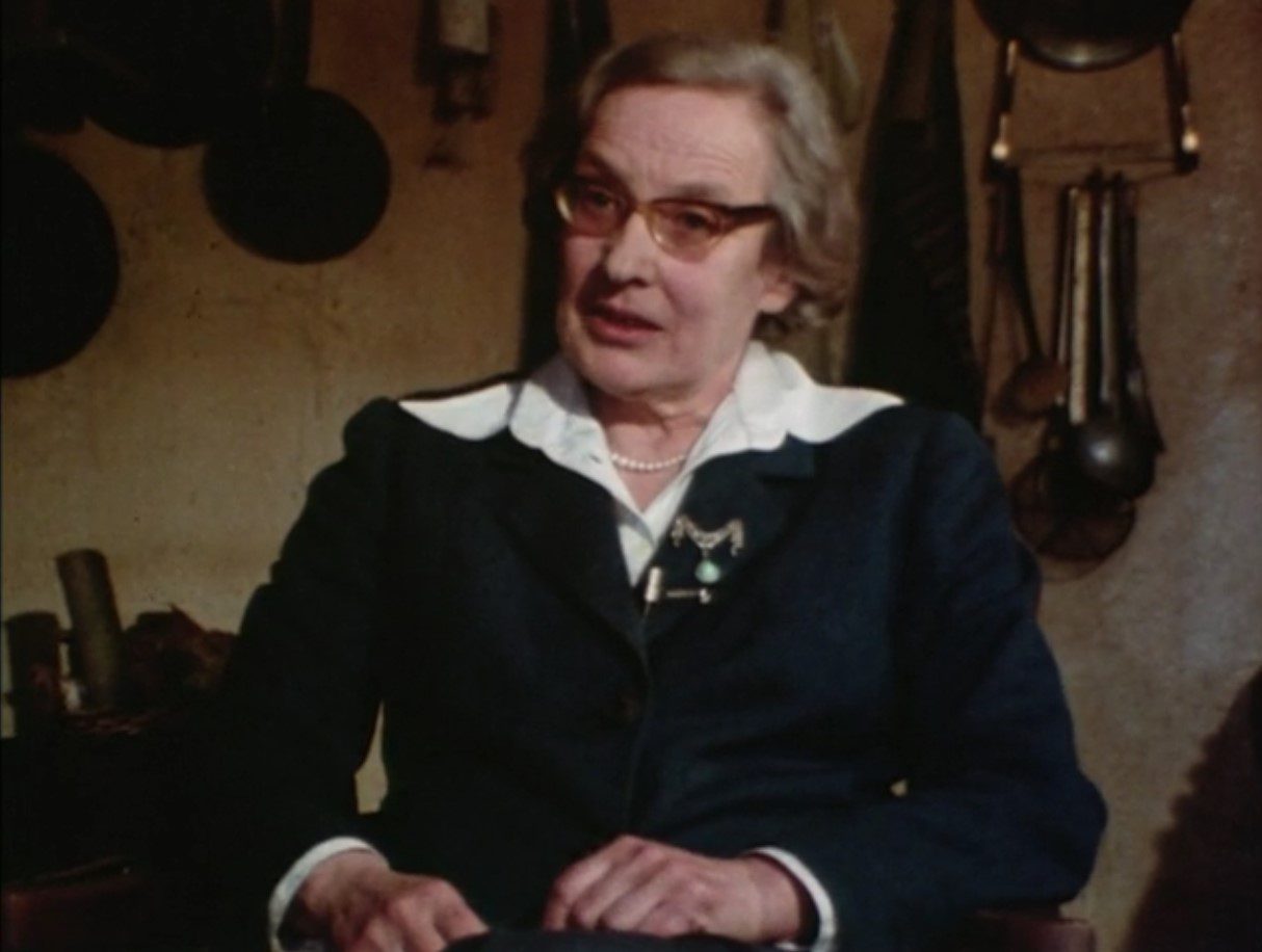 a woman with shorter silver hair wearing tinted glasses and a dark blazer sits with her fists resting in her lap.
