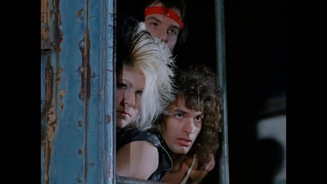 Three punks look out from behind a wooden building.