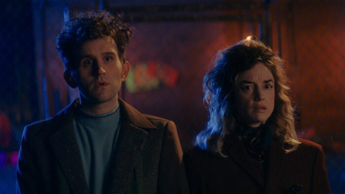 Harry Melling and Andrea Riseborough standing on the street at the beginning of the film watching The Young Gents kill a person on their stoop.