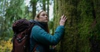 Wearing a backpack and jacket, Ruth (Maika Monroe), touches a tree on her venture into the woods. prepare for heir venture into the woods.