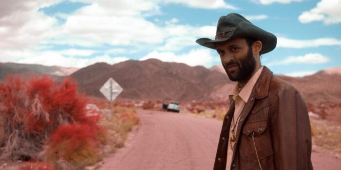 A bearded man in a cowboy hat stands alongside a desert road.