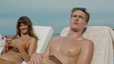 Charlbi Dean and Harris Dickinson lay on towel-covered pool chairs, in a pastel-colored still from 'Triangle of Sadness'