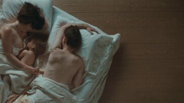 From left to right: Klara (Fides Krucker), a young Zoe (Magdalena Zokolowski) and a young Mitchell (James D. Watts) lie in bed together, partially undressed and covered up by the white bedsheets.
