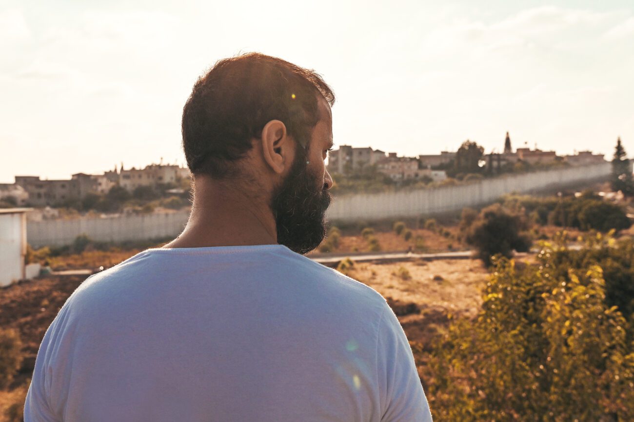 A bearded man (Mustafa, played by Ali Suliman) with back to the camera stares at the desert horizon.