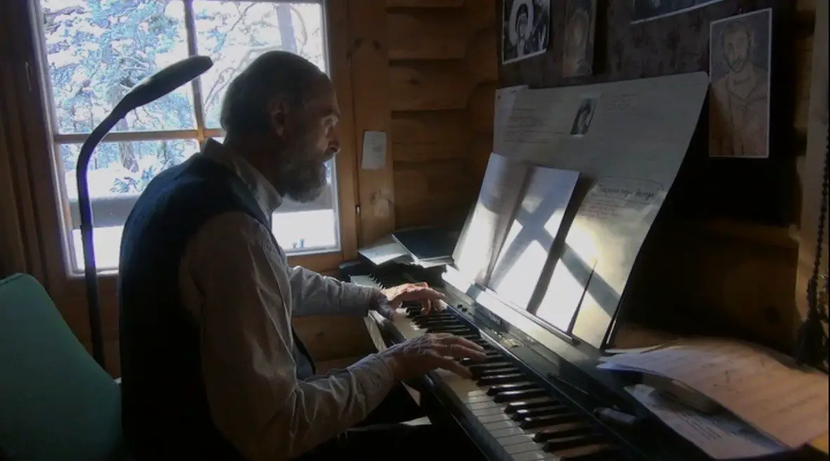 An older, balding man with a thick beard sits in front of a keyboard piano. He sits near a window that looks out into a forest.