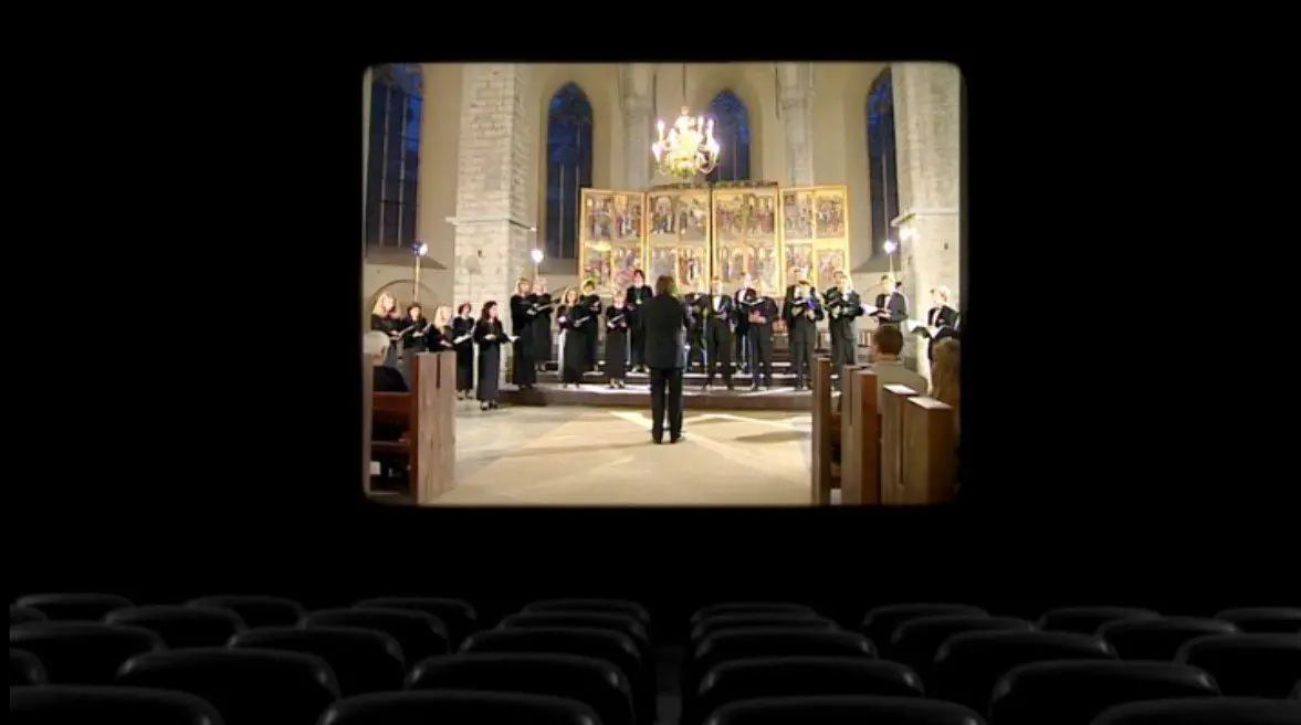 A movie screen floats above several empty theater seats. The screen features a brightly lit image of a choir dressed in black performing on the alter of a large cathedral.