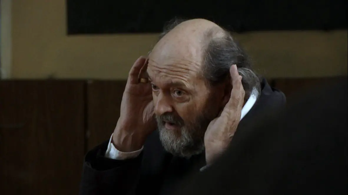 A balding man with a thick grey beard holds his hands near his ears as if he is trying to hear with more focus and clarity.