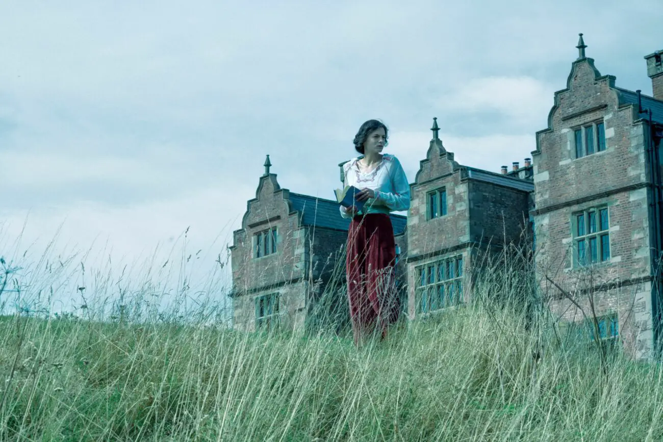 A woman stands with a book in an open field near a mansion in Lady Chatterley's Lover