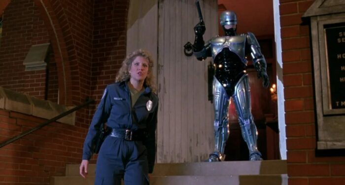 Lewis stands on stairs with Robocop, holding a gun, behind her.