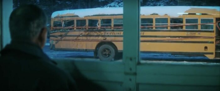 Image from The Sweet Hereafter: Mitchell Stevens (Ian Holm) stares at the ruined bus from beyond a window in a car repair shop.