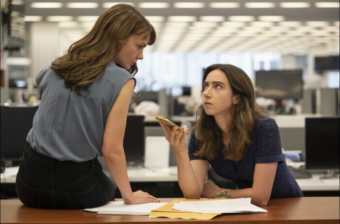 Megan Twohey (Carey Mulligan) sitting on a desk with Jodi Kantor (Zoe Kazan) sitting at the desk on speaker phone talking about the investigation