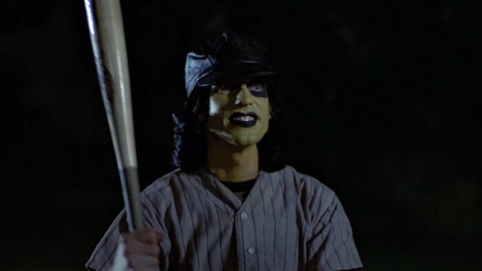 A gang member dressed like a baseball player with his face painted smiles while holding a bat
