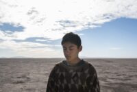 A young boy glances downward in front of an arid plain