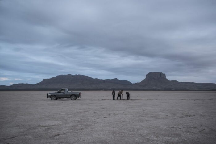 In a long shot on an arid desert plain, three men with shovels stand near a pickup.