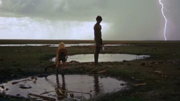 The native watches the boy look at a puddle with lightning behind them.
