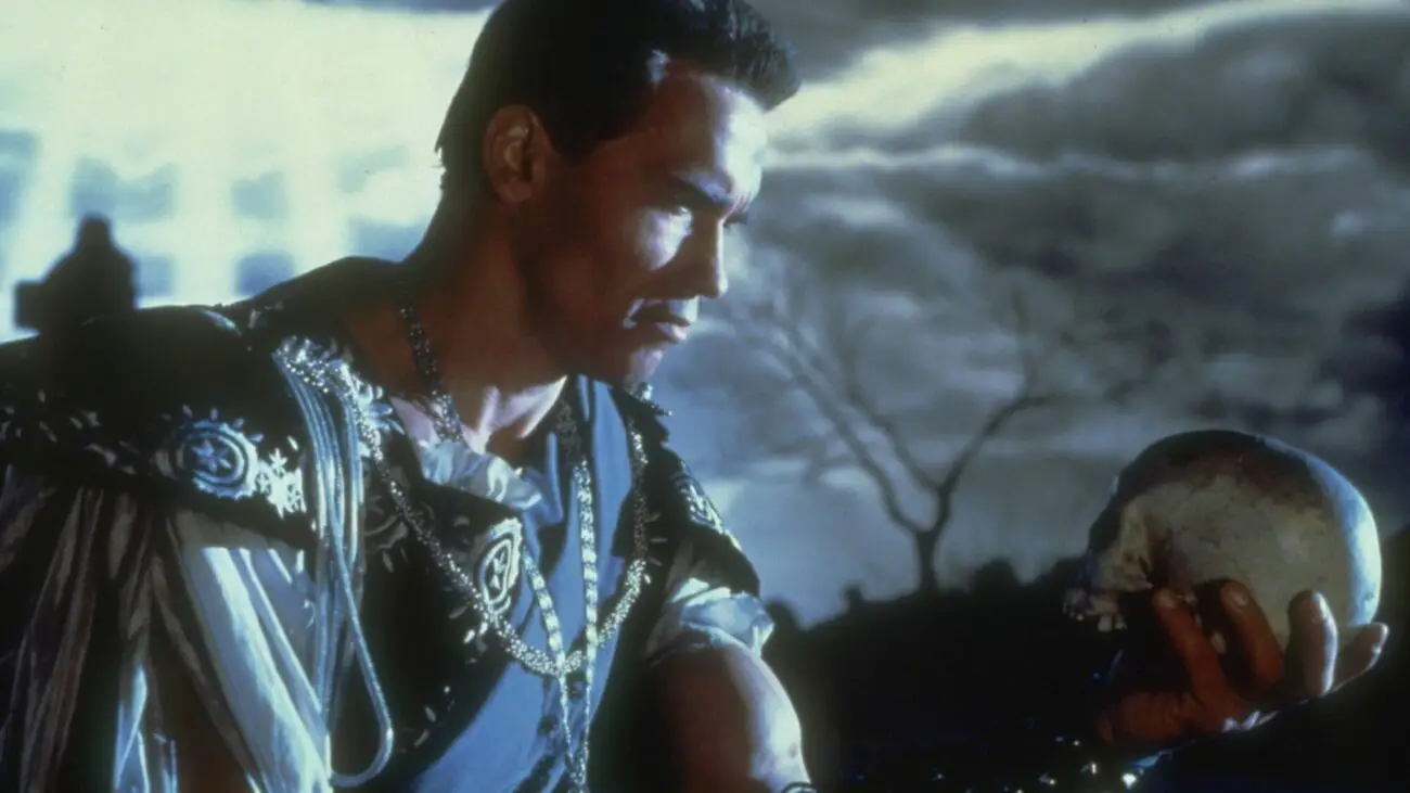 Arnols Schwarzenegger, as Hamlet, seated and looking at a skull he's holding in his left hand.