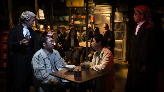 Mak Pui-tung as Angus and Alan Yeung as Henry sit in Henry's apartment with barristers and jury members observing.