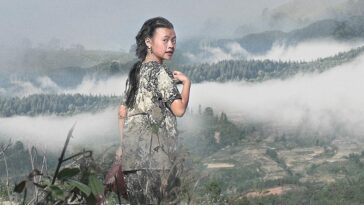 A Hmong girl looks back at the camera, behind her a landscape of mountains enshrouded in mist.