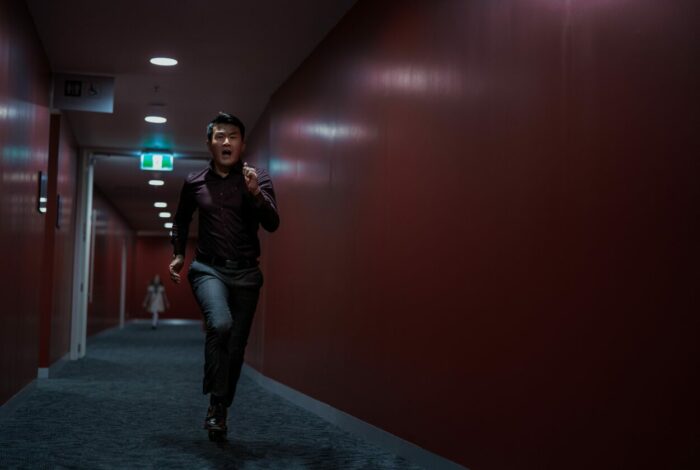David (Ronny Chieng) runs down a red-walled hallway from M3GAN.