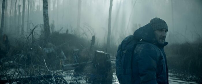 Mathieu, wearing a stocking cap, parka, and backpack, makes his way through a swamp.