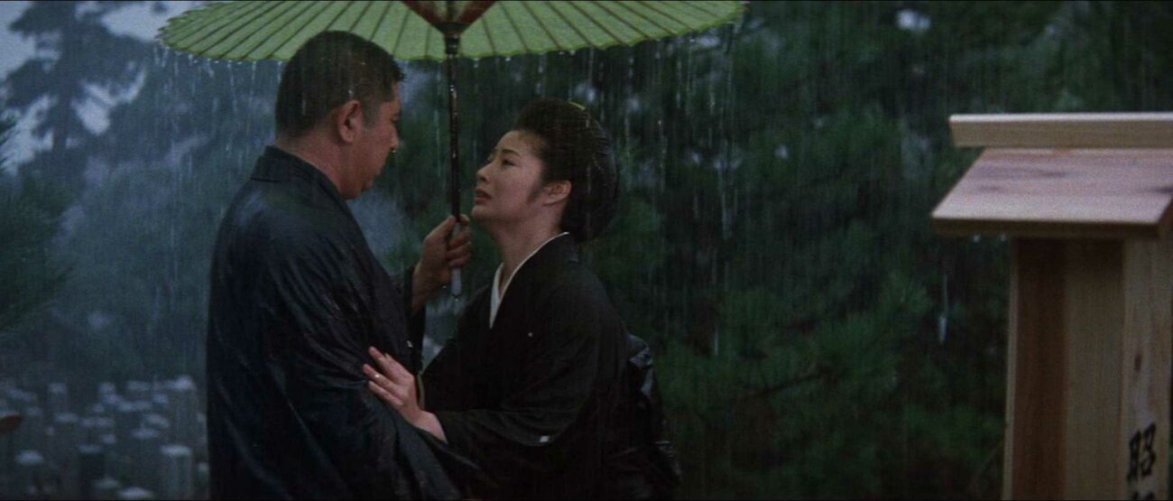Matsuda holds an umbrella while standing in the rain as his wife holds him, looking upset.
