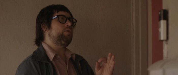 Clay Tatum playing Clay in The Civil Dead, wearing thick black framed glasses, bearded, and burdened with a bad haircut.