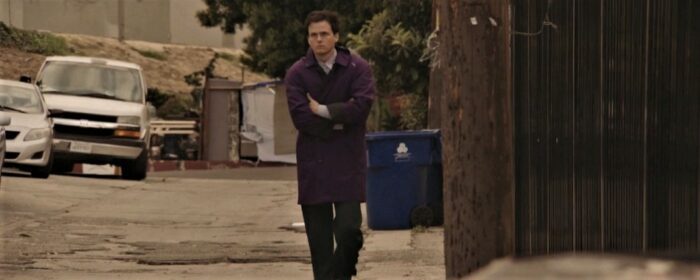 Whitmer Thomas as Whit in The Civil Dead, walking alone down a deserted city street, wrapped in a purple long coat.