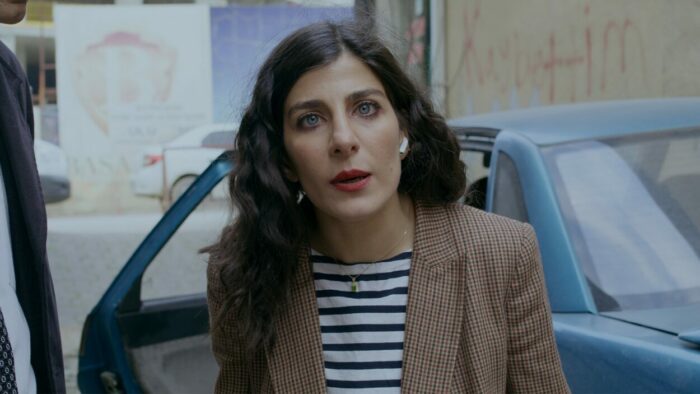An Iranian woman dressed in a blouse and blazer exits a car and addresses the camera directly.