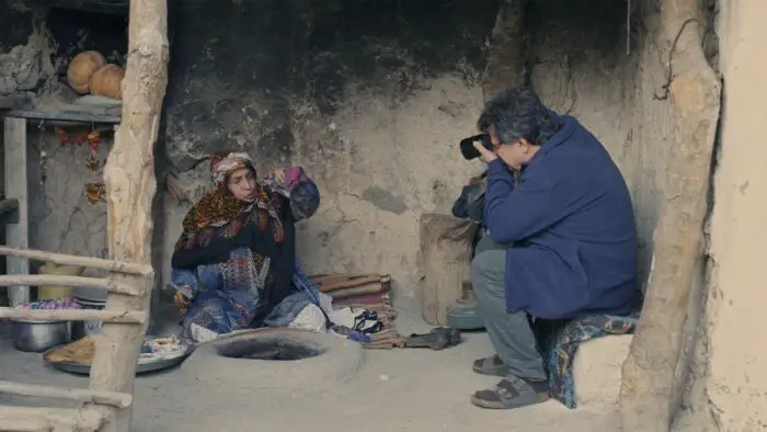 A man in a blue jacket photographs a Turkish woman as she prepares a meal.