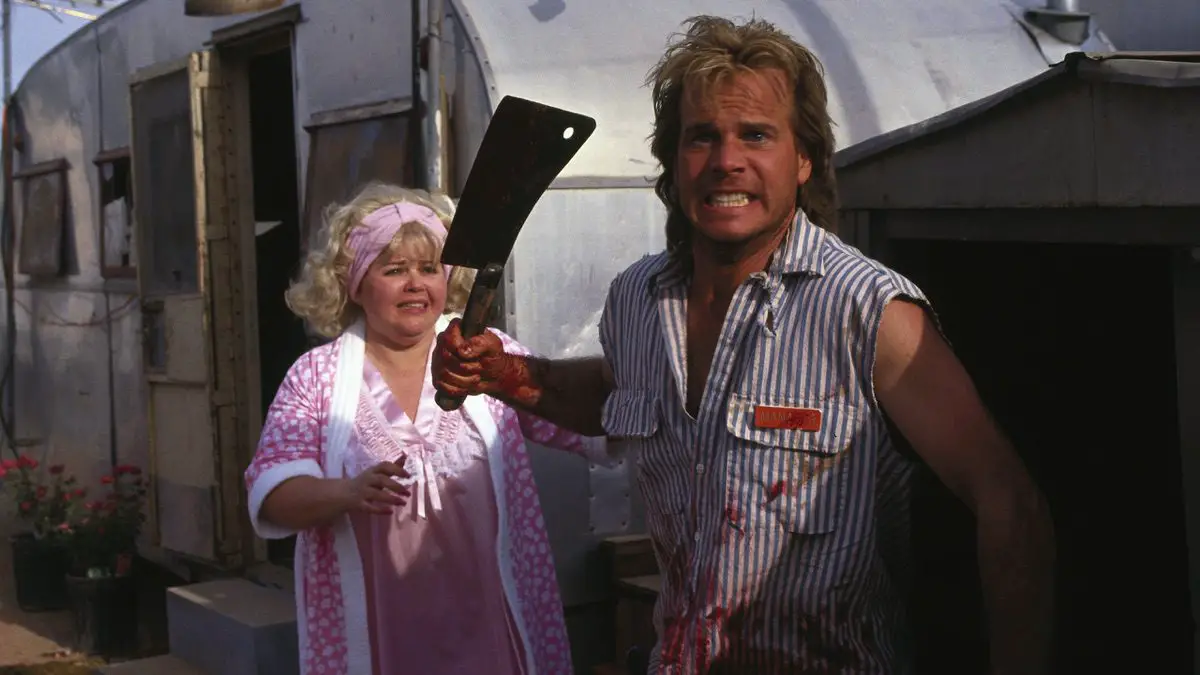 Graham stands in front of a trailer, holds up a meat cleaver, angry, with Doattie standing behind him.