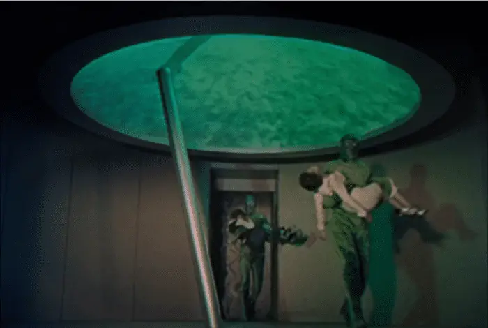 Green Aliens carry a woman and a child inside a green space ship with a metal rod sticking up from the floor 