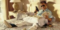 A man dressed in a Hawaiian shirt sits on a hotel bed brandishing a gun.