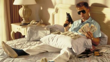 A man dressed in a Hawaiian shirt sits on a hotel bed brandishing a gun.