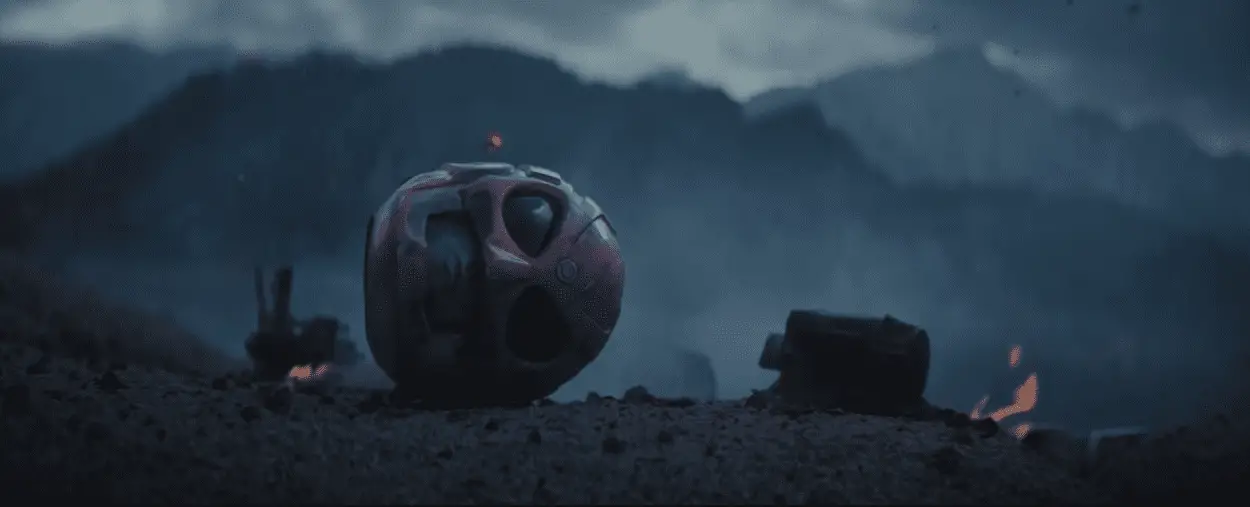 The Pink Power Ranger helmet lays sideways on the ground, next to a burning ember, with a smoky vista of mountains behind it.