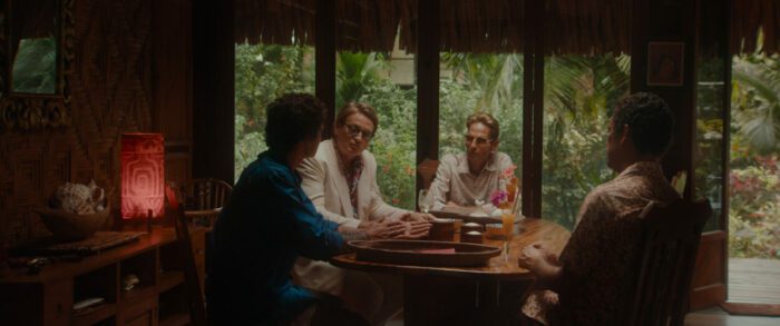 Four men sit at a table in a tropical hut.