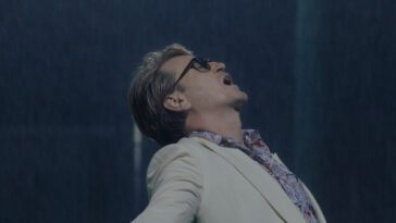 A man wearing a white sport coat, sunglasses, and a tropical shirt opens his arms as rain falls.