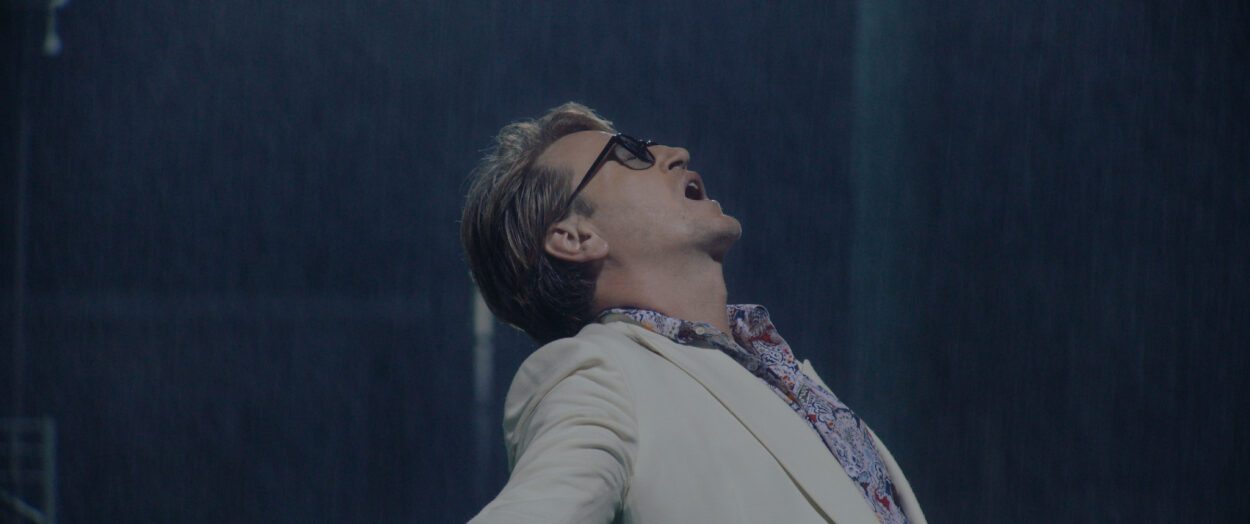 A man wearing a white sport coat, sunglasses, and a tropical shirt opens his arms as rain falls.