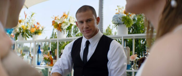 Mike (Channing Tatum), wearing a waiter's vest and black tie, prepares drinks as a bartender for a couple.