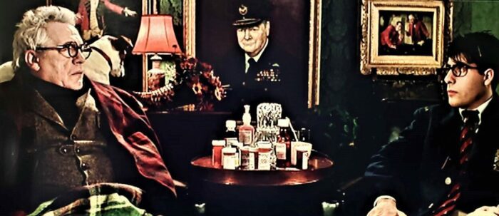 Brian Cox as Dr. Guggenheim, headmaster of Rushmore Academy, addressing Max Fischer (Jason Schwartzman). The two sit in leather chairs with a stack of medicine on a table between them, a frame oil painting of Winston Churchill in the background between them.