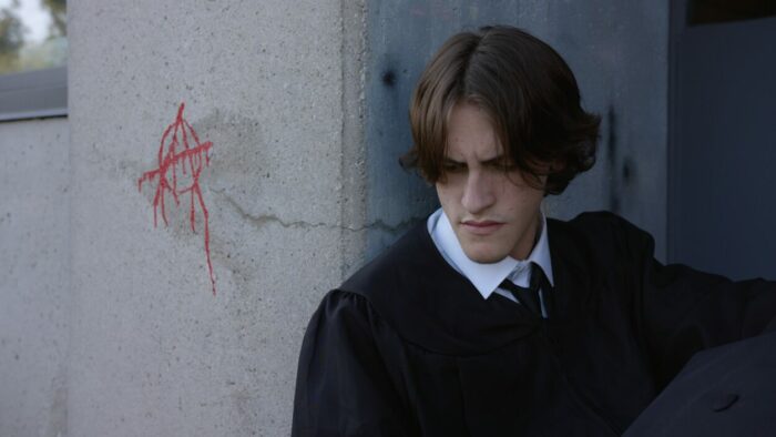 A boy in his graduation gown rests against a concrete wall.