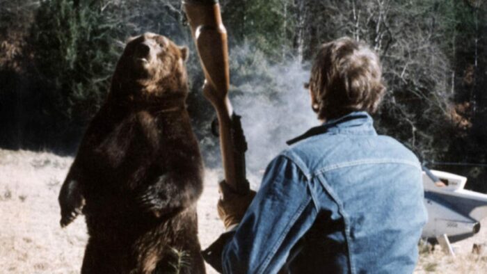 The bear stands on its hind legs with Stober in the foreground, his back to the camera, holding a rifle.