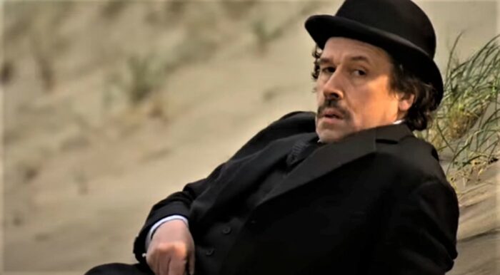 Stephen Rea as Leopold Bloom in the 2003 adaptation of Ulysses by James Joyce. Bloom lounges in a black suit on the beach while wearing a bowler hat.