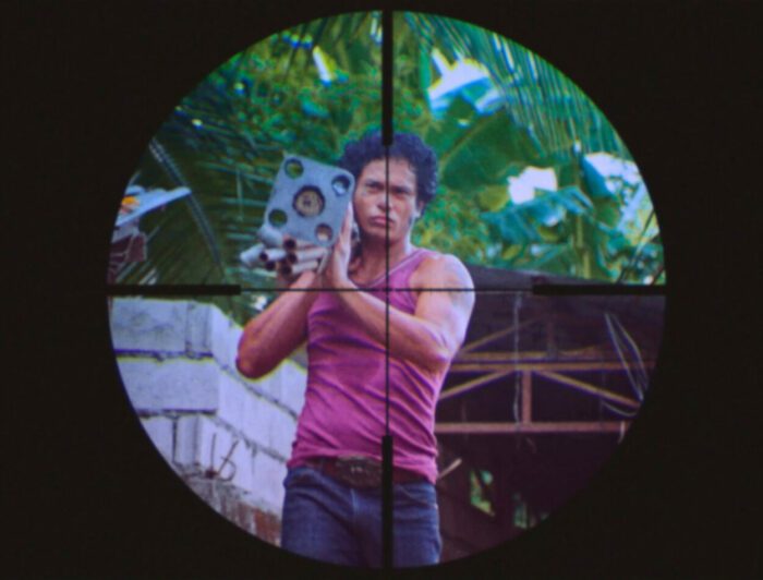 A man is depicted carrying a weapon through the crosshairs of a rifle scope.