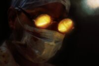 The Surgeon with a mask over his face and glasses on looks down. The glasses reflect fire in the lenses.