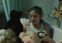 Giraffe (Maria Abreu) in her room, surrounded by stuffed animals.