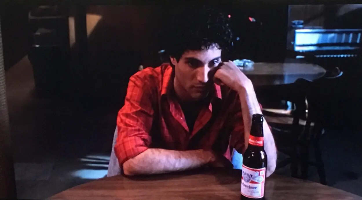 Ed Jr. sits at a table resting his head on his hand with a beer on the table in front of him.
